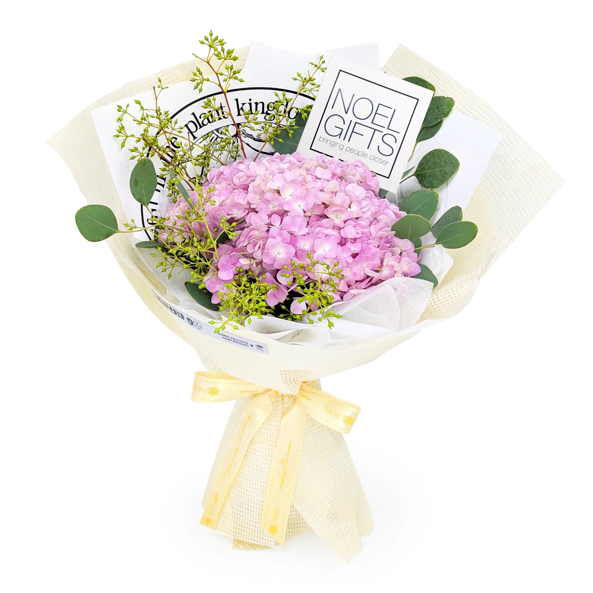 A wrapped flower bouquet featuring pink hydrangeas, green eucalyptus leaves, and small yellow-green filler stems, tied with a pale yellow ribbon. Cardboard backing and printed inserts are visible behind the flowers, including the text 'NOEL GIFTS' and 'The Plant King'.