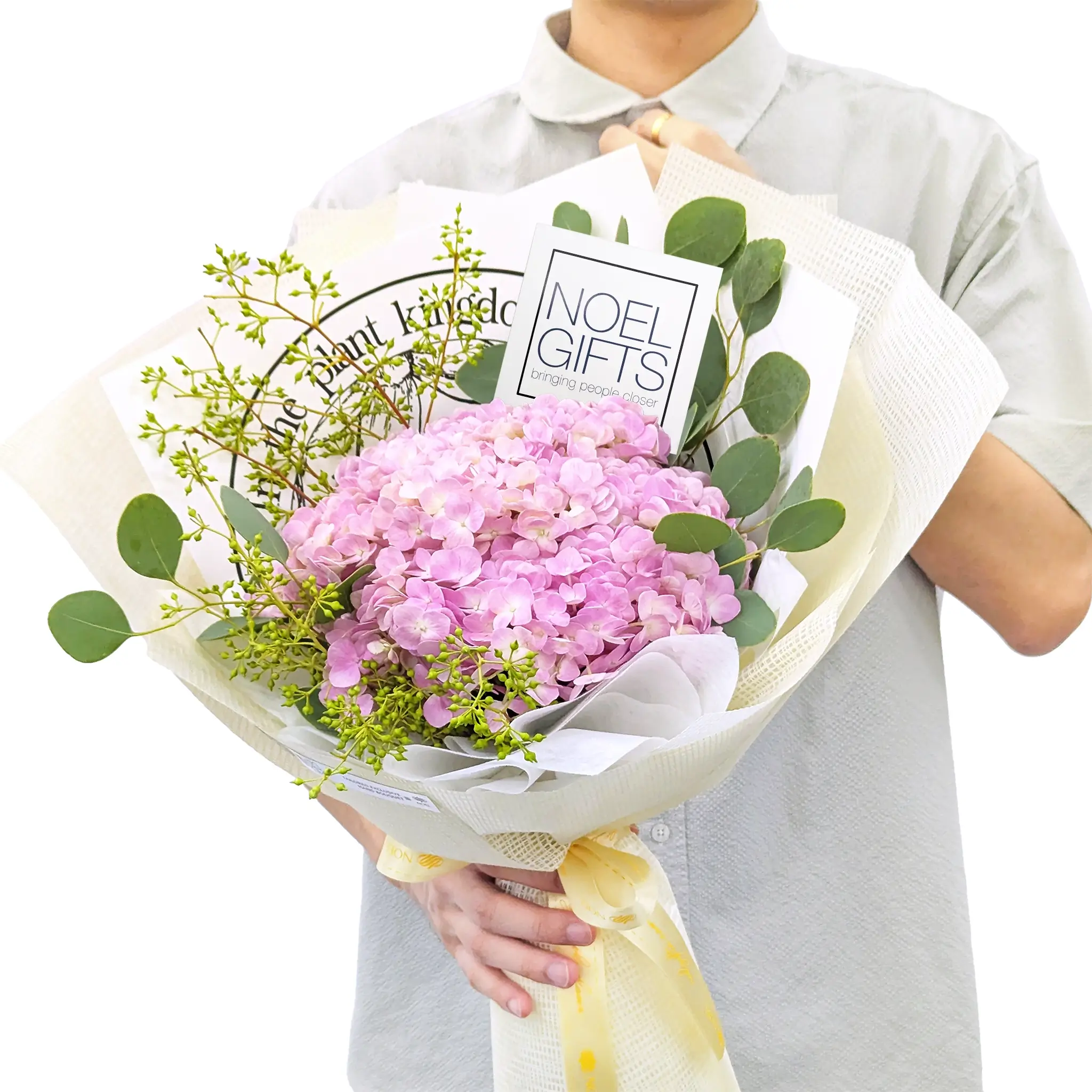 A person holding a wrapped bouquet of pink hydrangeas with green foliage and eucalyptus stems. The bouquet is wrapped in cream paper with a yellow ribbon, and a card reading 'Noel Gifts' is visible inside.