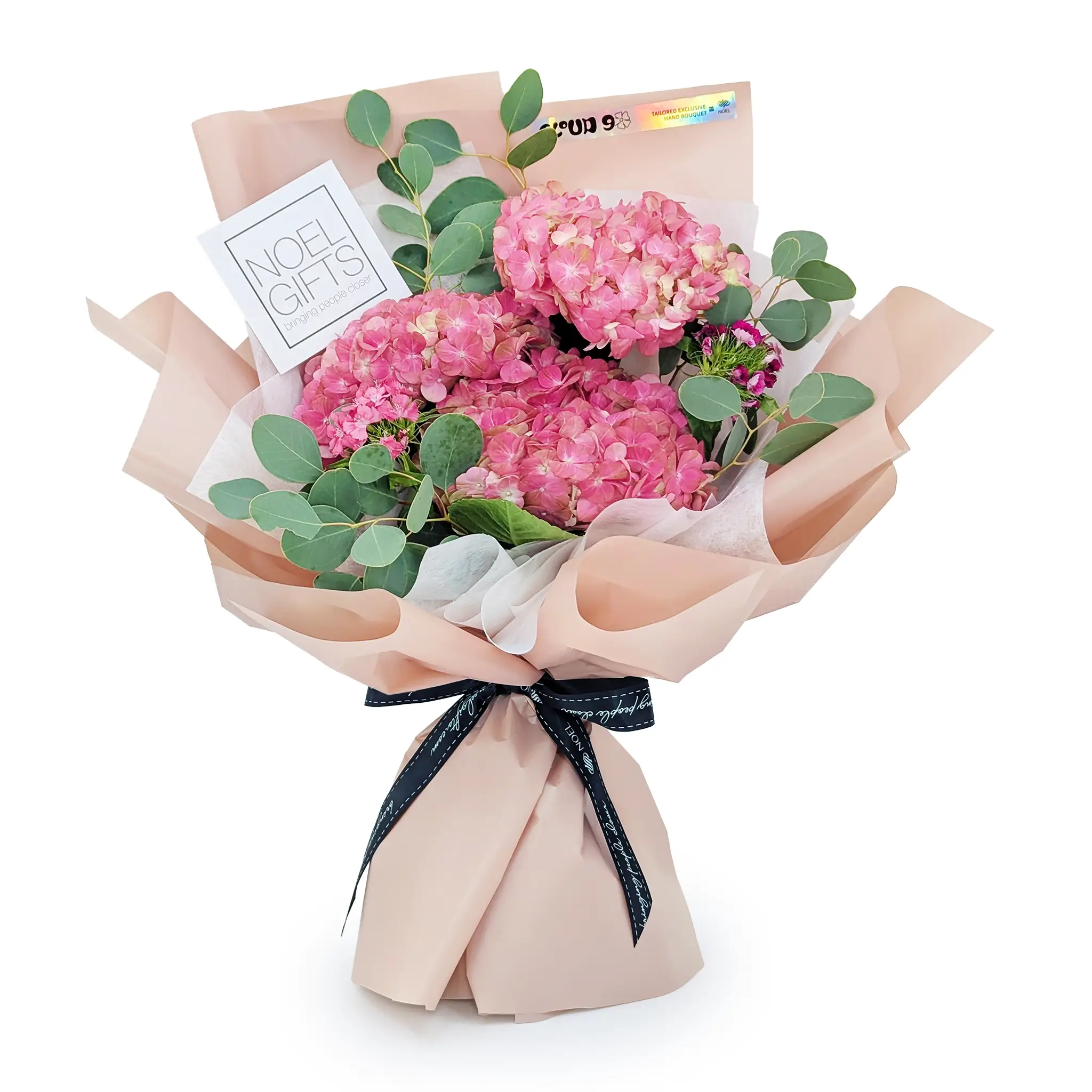 A bouquet of pink hydrangeas wrapped in blush pink tissue and kraft paper, with green eucalyptus stems, tied with a black ribbon, and a white gift card reading 'NOEL GIFTS' on a clean white background.
