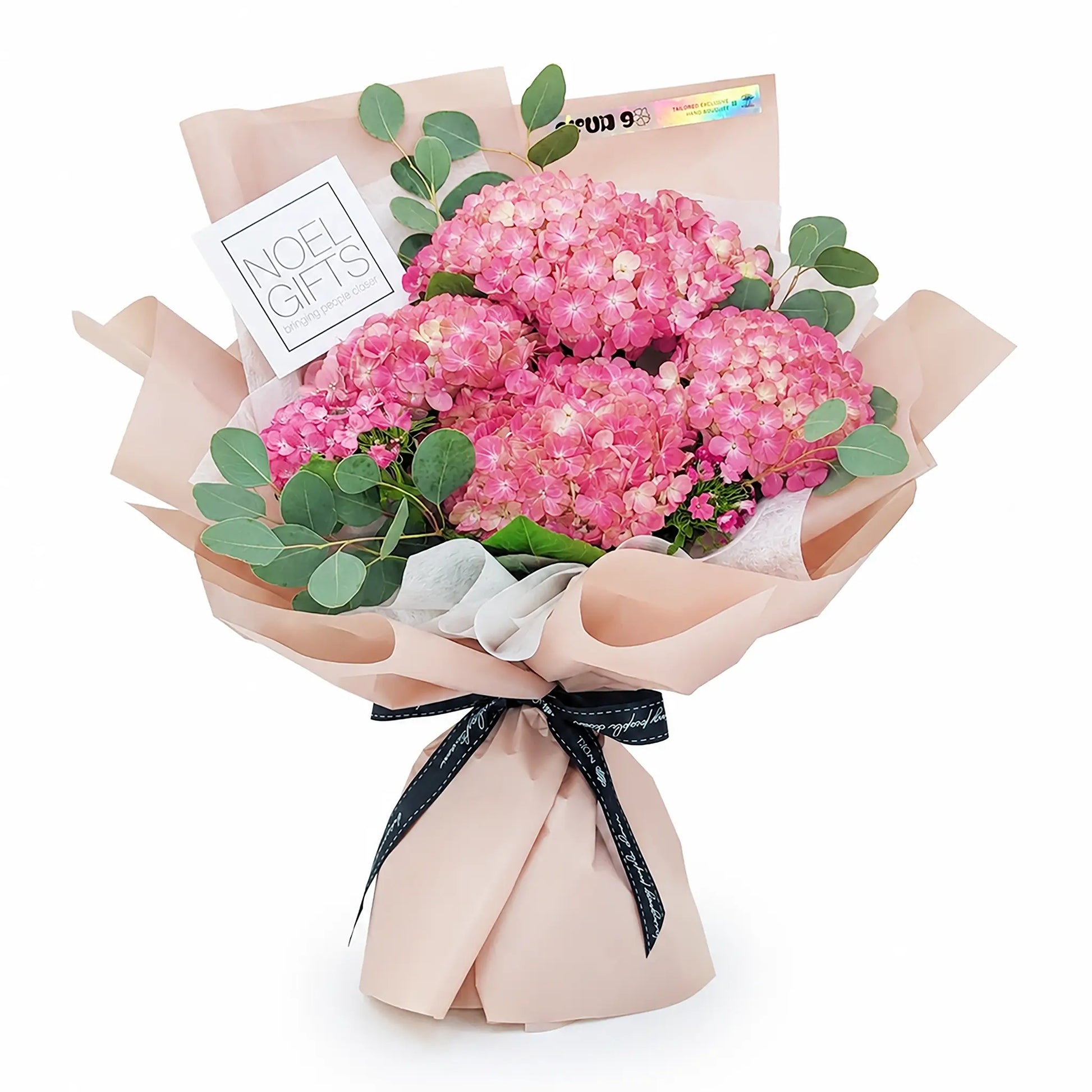 A bouquet of pink hydrangeas wrapped in blush pink paper with a black ribbon, a small white card reading 'NOEL GIFTS', and green eucalyptus leaves, photographed on a white background.