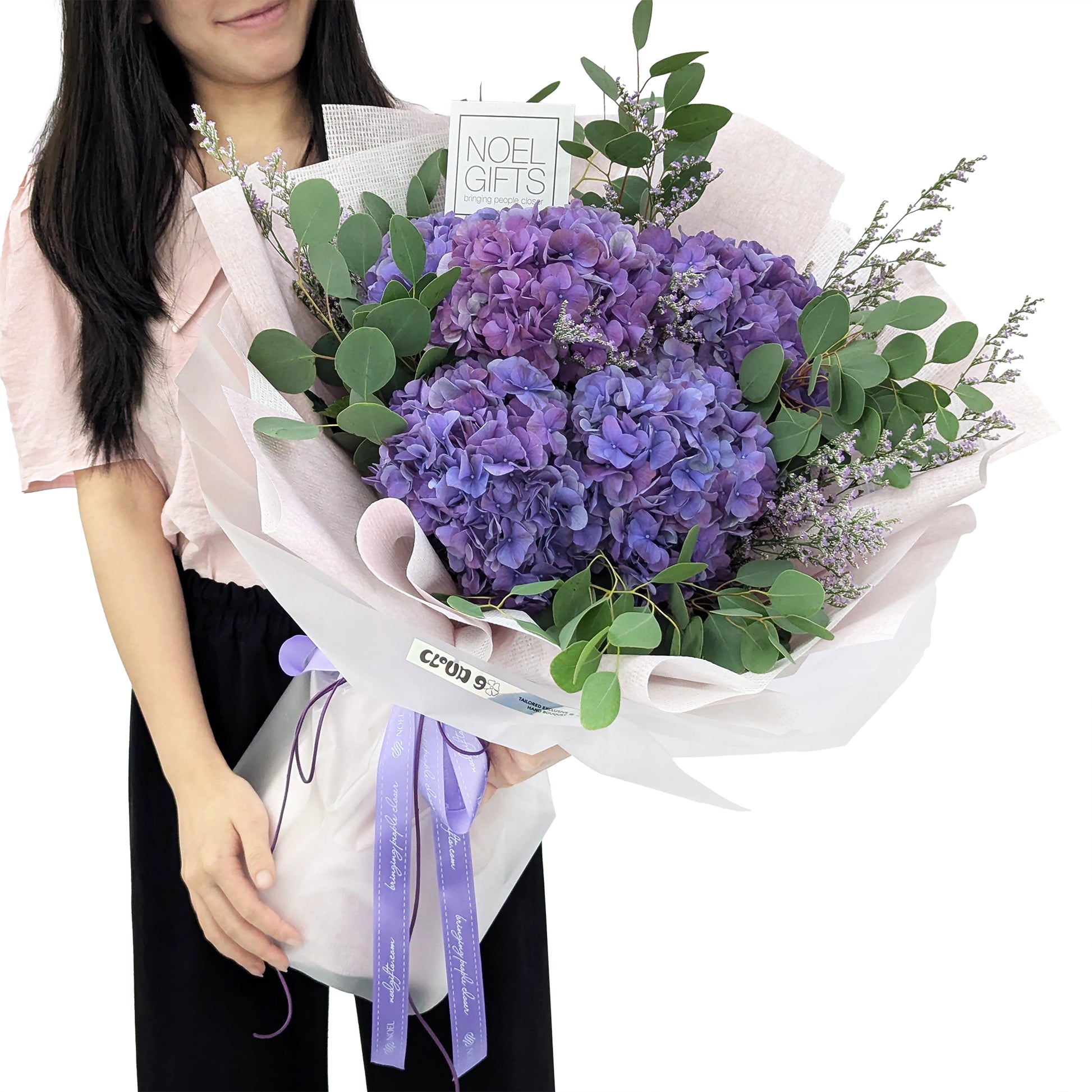 A person holding a large floral bouquet wrapped in pale pink tissue paper, with purple hydrangeas as the main flowers and green foliage. A small card reads 'NOEL GIFTS' and the bouquet has lavender ribbon ties.