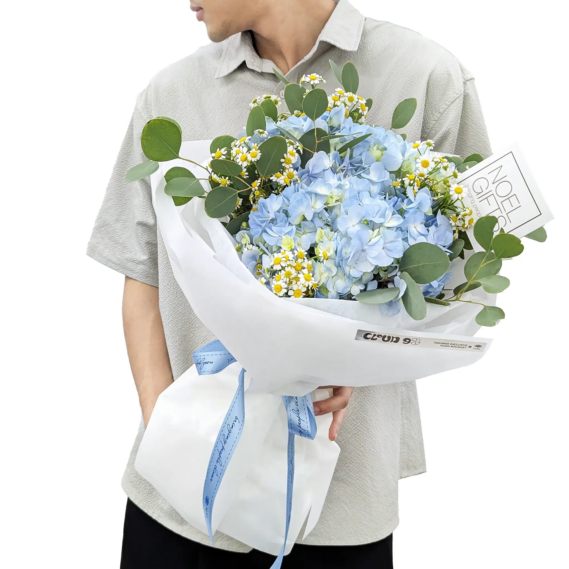 A person holding a wrapped bouquet of light blue hydrangeas and small white/yellow daisies with green eucalyptus leaves. The bouquet is wrapped in white paper with a blue ribbon, and a small card is tucked into the arrangement.
