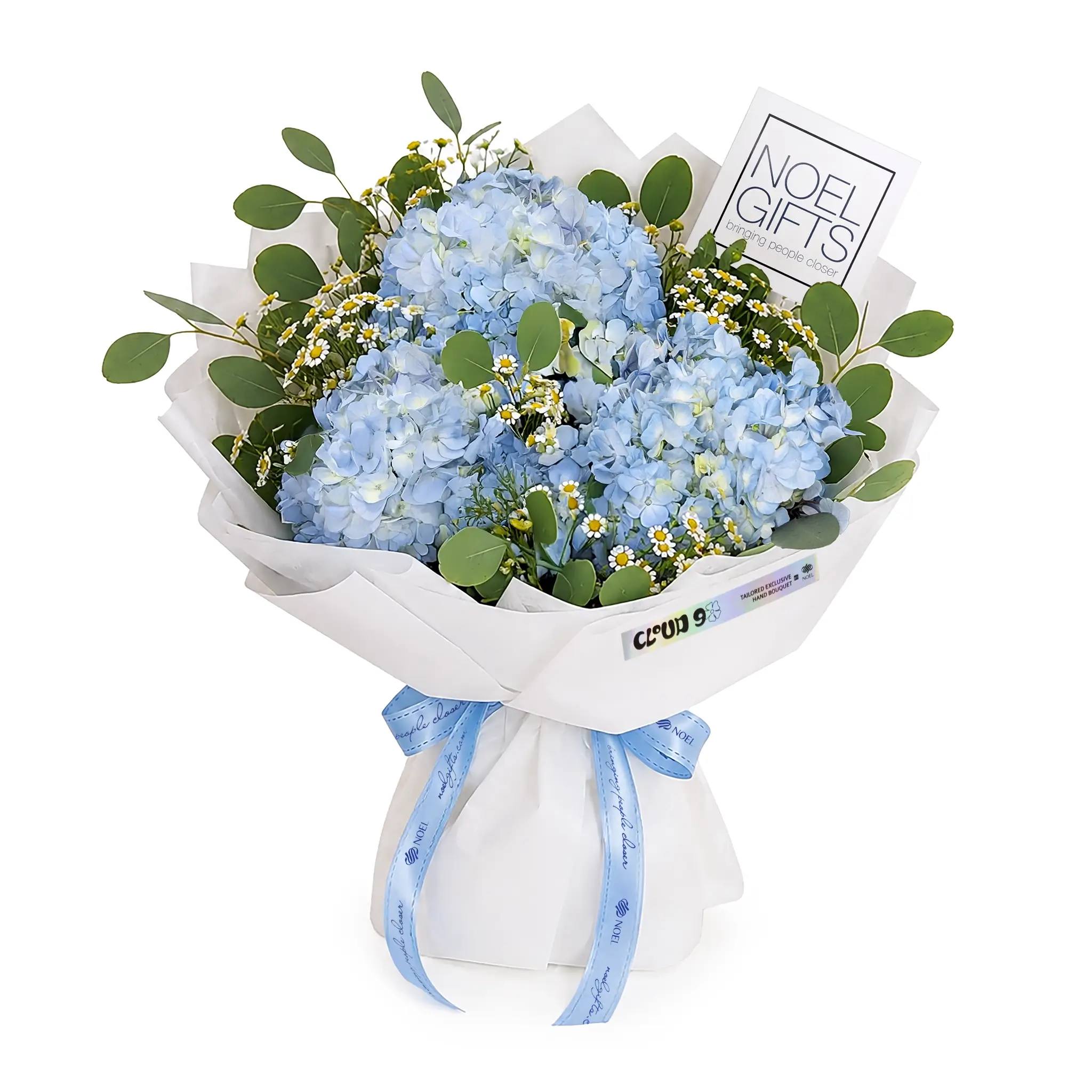 A wrapped floral bouquet featuring light blue hydrangeas, green eucalyptus leaves, and small white daisies, tied with a light blue ribbon. A white card reading 'NOEL GIFTS' is tucked into the bouquet, and the package is wrapped in white paper with a matching ribbon.