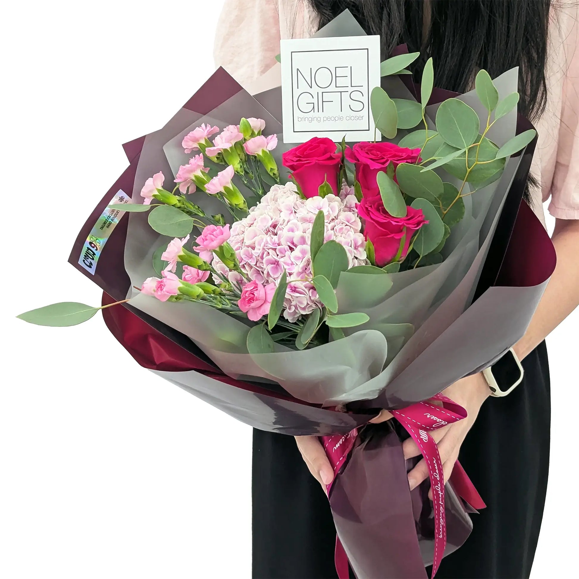 Person holding a wrapped bouquet of pink carnations, light pink hydrangeas, red roses, and eucalyptus leaves, with a “NOEL GIFTS” card.