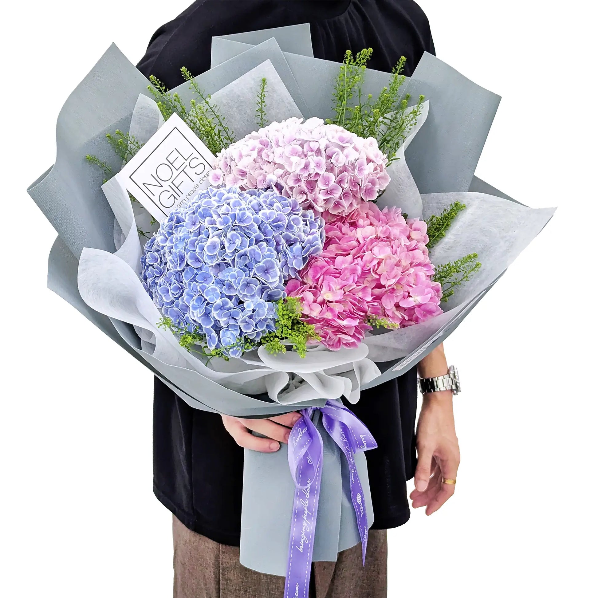 A bouquet of four hydrangea flowers in pastel blue, lavender, pink, and white, wrapped in light gray paper with a purple ribbon and a card reading 'NOEL GIFT'. A person is holding the bouquet against a plain white background.