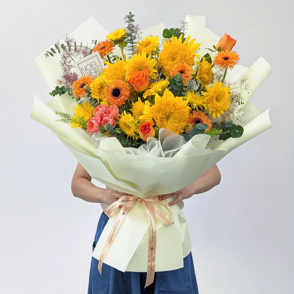 A vibrant flower bouquet held by a person against a plain light grey background. The bouquet features a mix of yellow Teddy Bear Sunflowers, orange gerbera daisies, pink roses, and green foliage, wrapped in colored paper with a beige ribbon.
