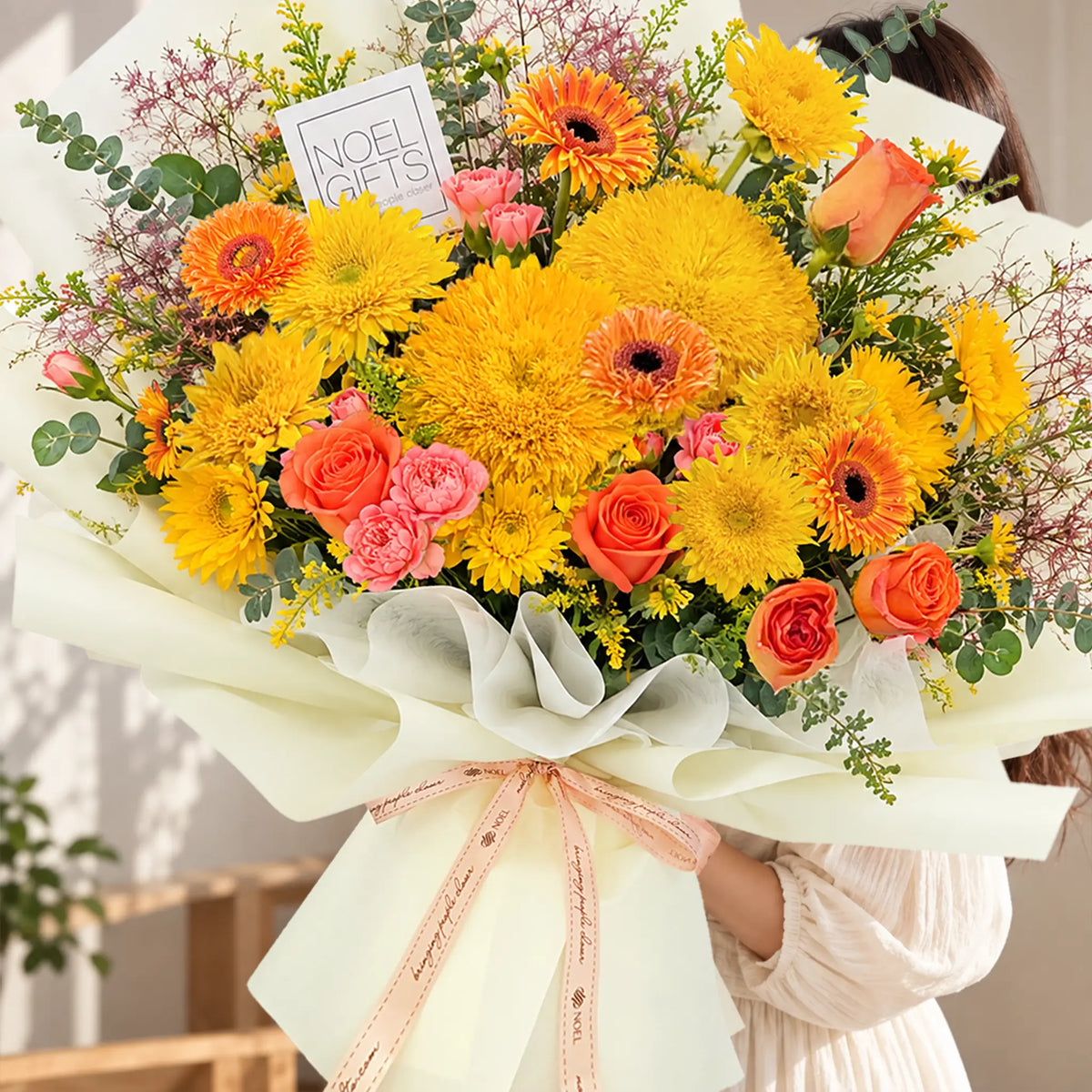 A large, vibrant floral bouquet wrapped in cream paper and tied with a pink ribbon, held by a person wearing a white long-sleeve top. The bouquet features a mix of bright yellow Teddy Bear Sunflowers, orange gerbera daisies, pink roses, and green foliage, with a small card that reads 'NOEL GIFT'.