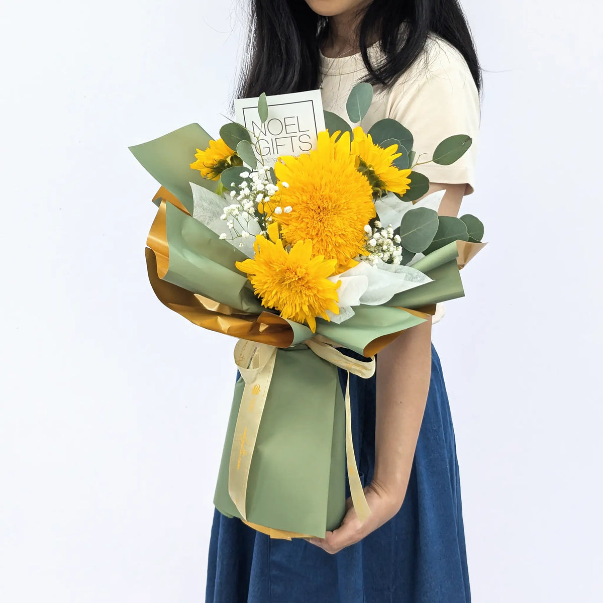 A person holding a wrapped flower bouquet with bright yellow flowers, green foliage, and sage green paper wrapping with gold ribbon, against a plain light background.