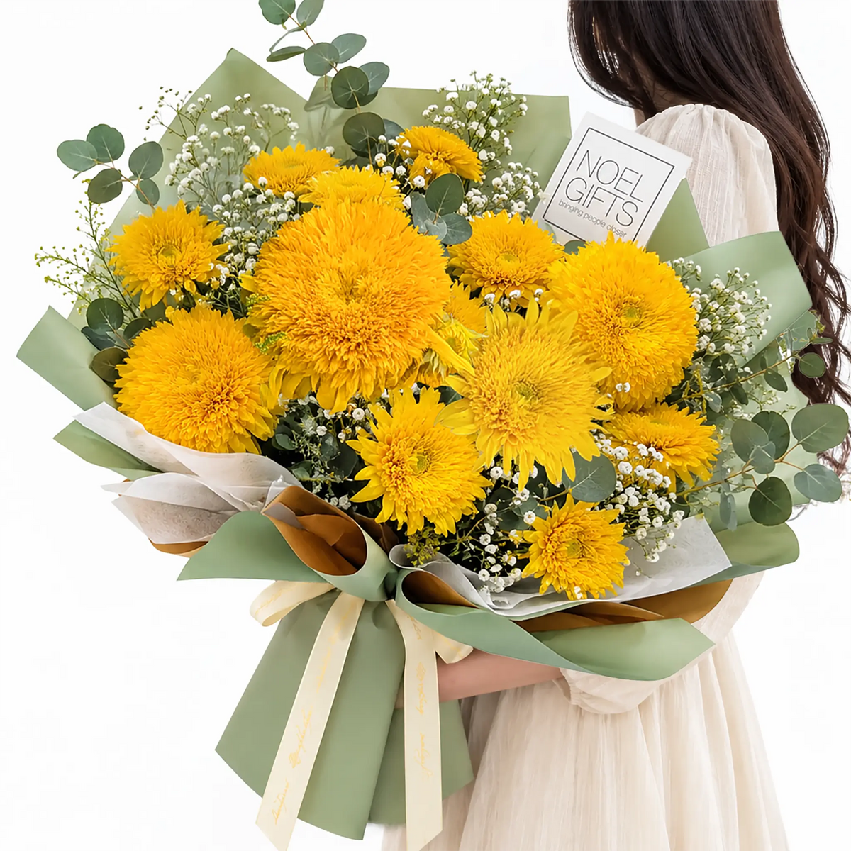 A large, vibrant yellow flower bouquet wrapped in sage green paper and tied with a cream ribbon, held by a person wearing a cream-colored dress against a solid white background. A small gift card reading 'NOEL GIFTS' is tucked into the bouquet.