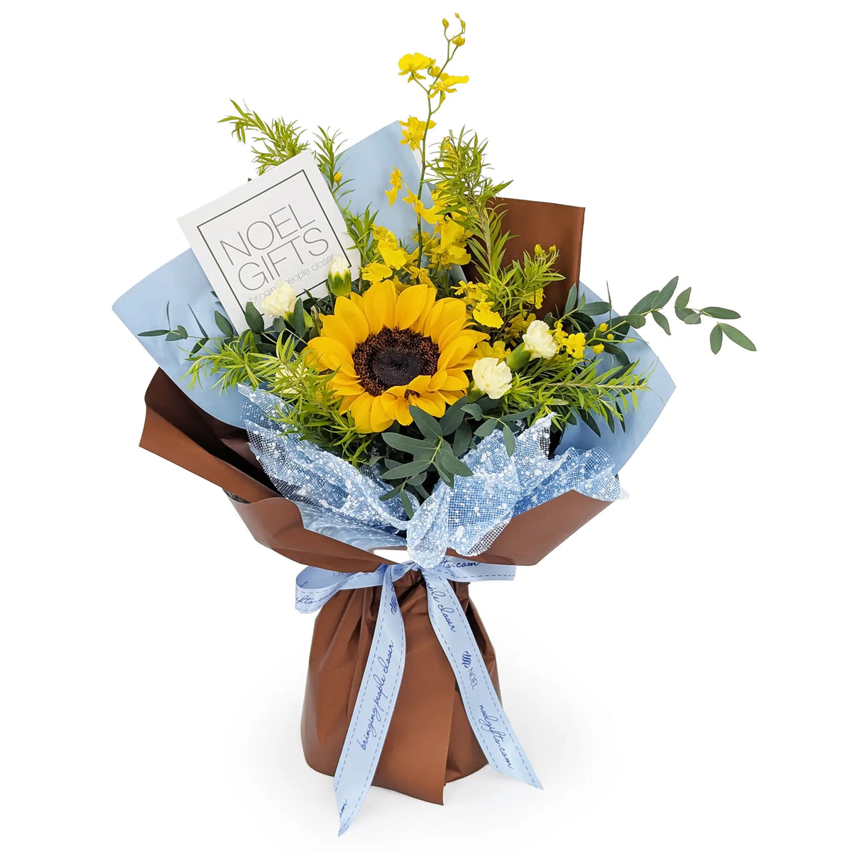 A floral gift bouquet featuring a large yellow sunflower, small white flowers, green foliage, and yellow blossoms, wrapped in brown paper with light blue tissue paper, a blue glittery ribbon, and a white card that reads 'NOEL GIFTS'.
