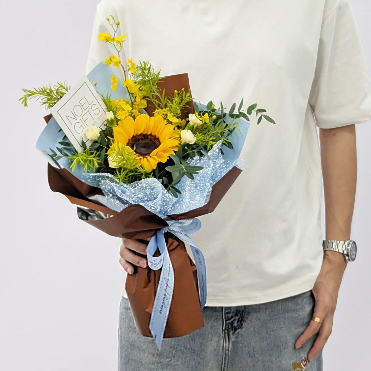 Blue Skies Petite Hand Bouquet featuring a yellow sunflower, yellow and white flowers, fresh greenery, brown wrapping, and a light blue ribbon with a Noel Gifts card.
