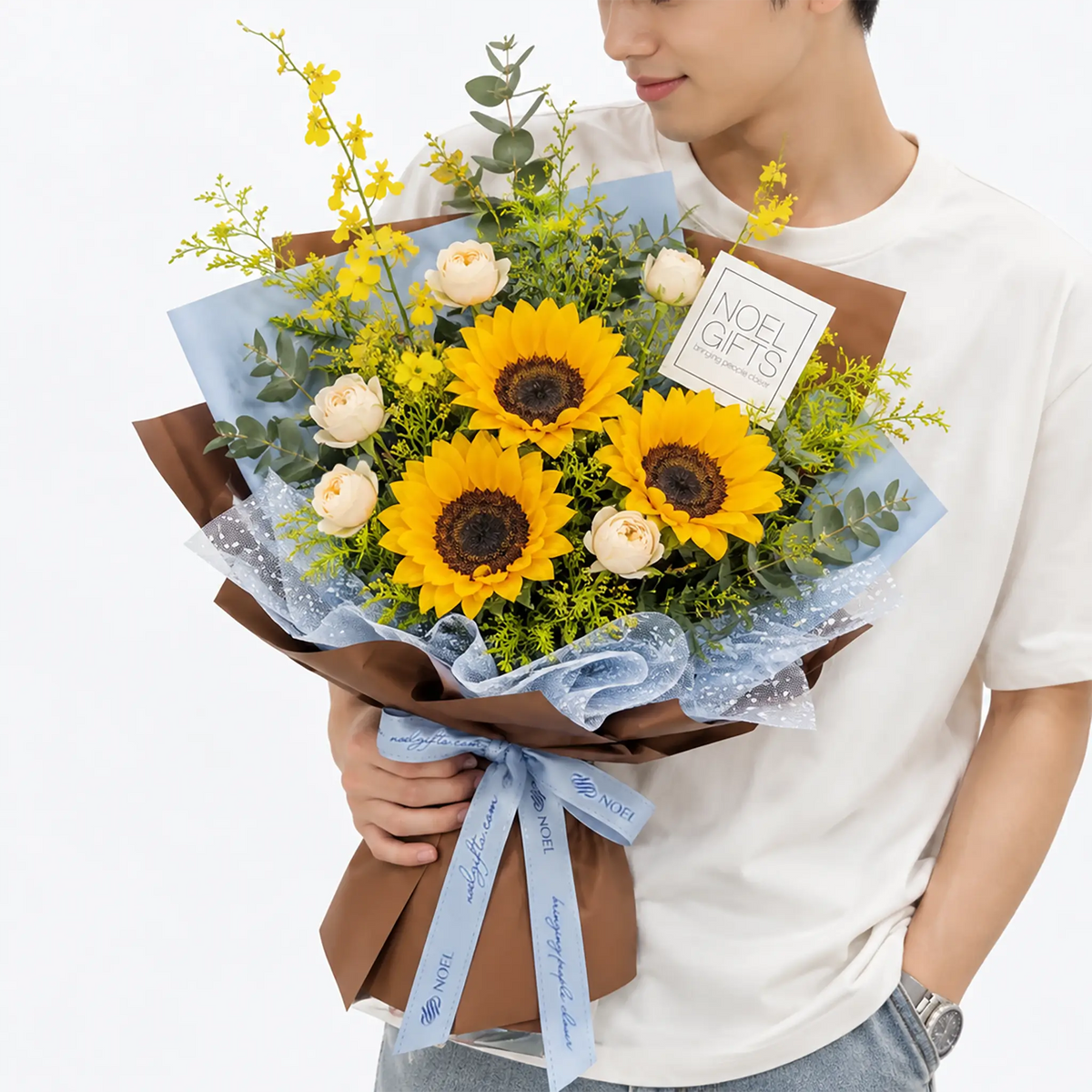 Man in a white T-shirt holding the Blue Skies Classic Hand Bouquet featuring 3 sunflowers, champagne spray roses, yellow Oncidium orchids, Callistemon hybridus and eucalyptus foliage in blue and warm brown wrapping with a light blue ribbon | Noel Gifts